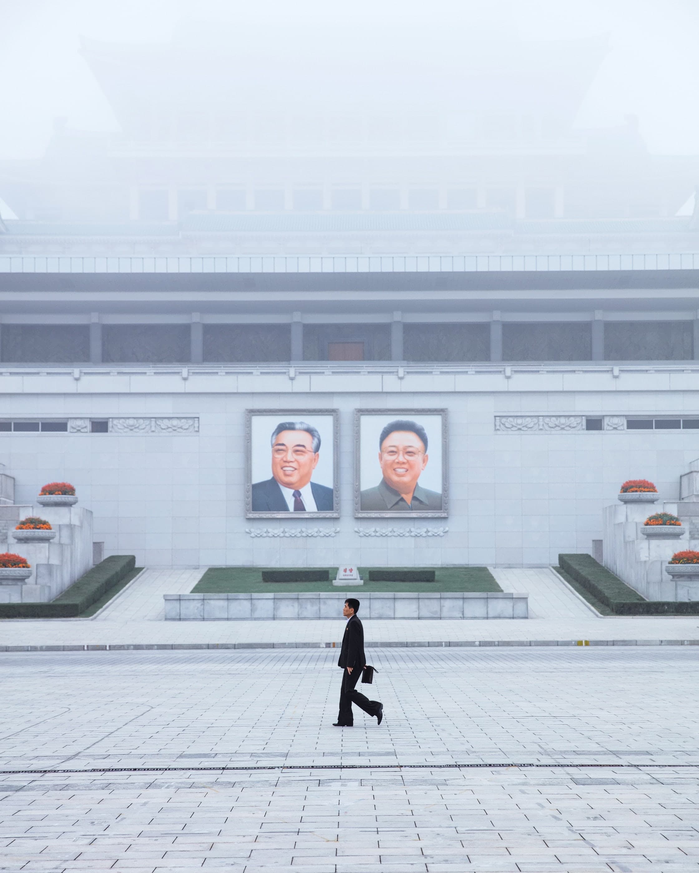 A suited man crossing a vast plaza beneath two leader portraits, Pyongyang